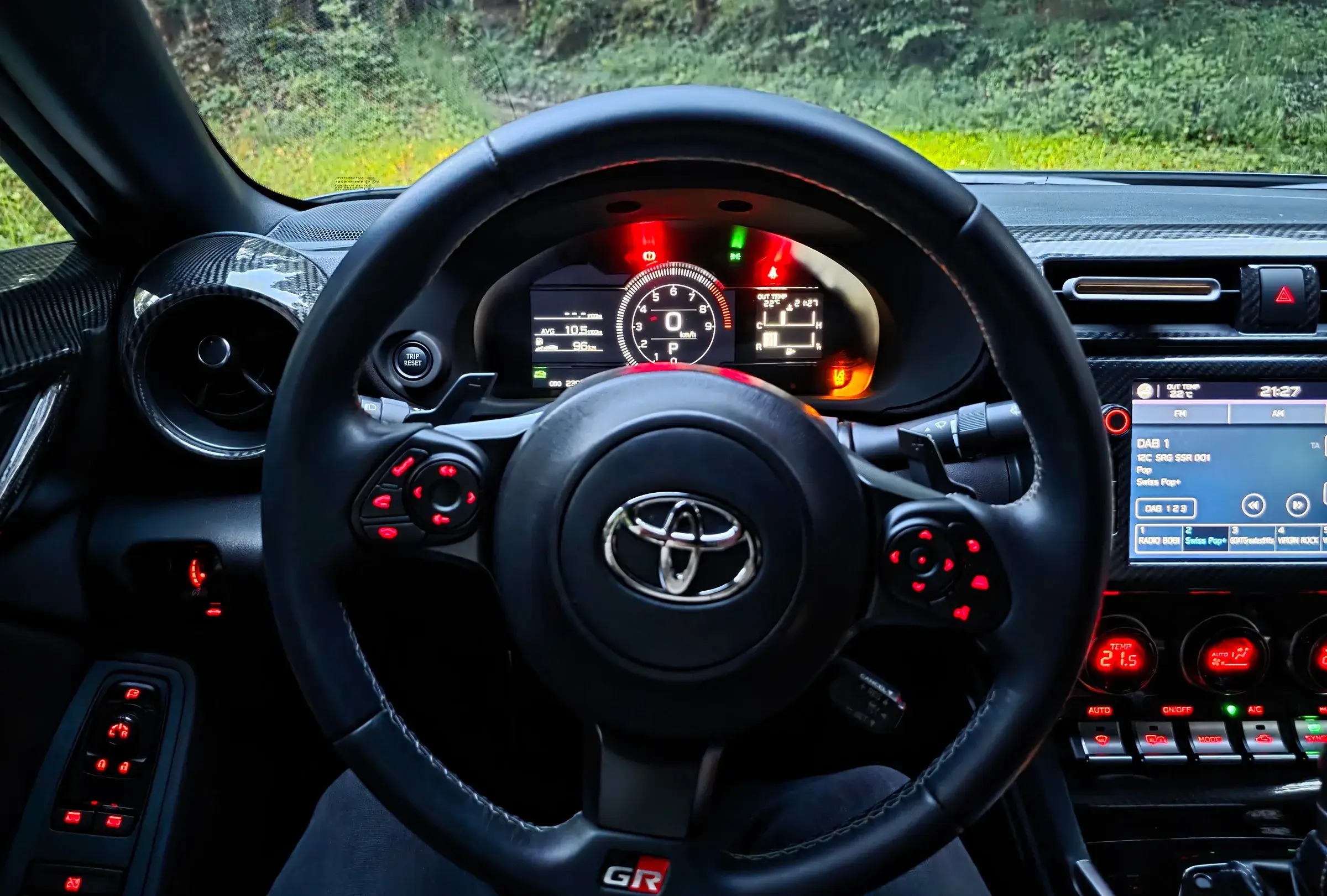 Cockpit des Toyota GR86 aus der Sicht des Fahrers am Abend: Lenkrad, beleuchtete Instrumente, dahinter der Waldrand