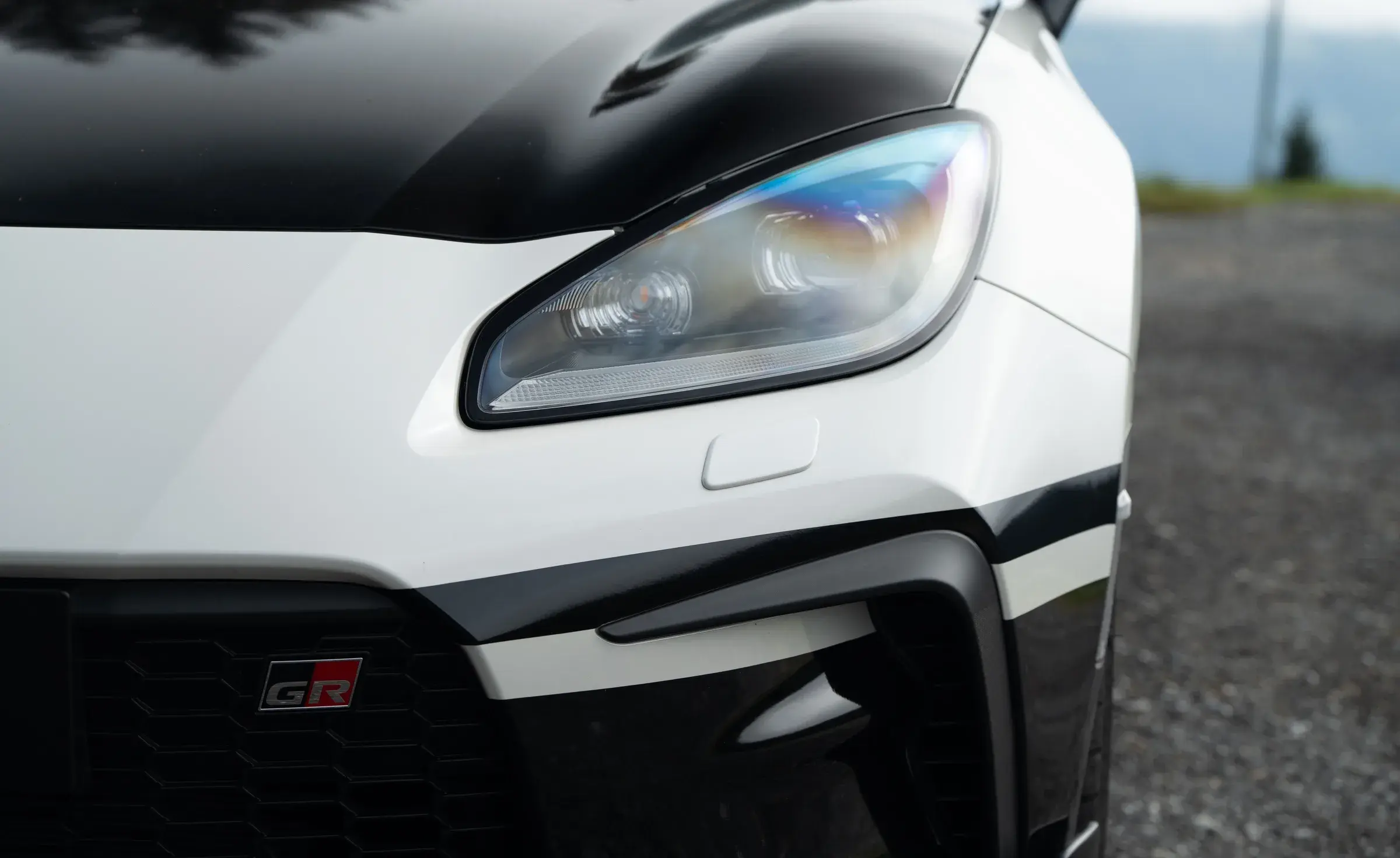 Headlight and front corner of the Initial Drive Toyota GR86 in daylight, with the GR badge on the grille