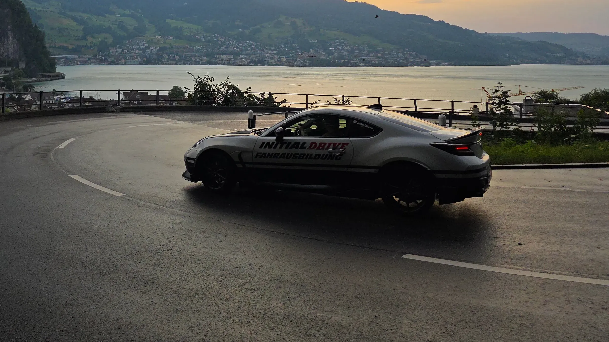 Toyota GR86 von Initial Drive fährt am Abend durch eine Haarnadelkurve über dem See