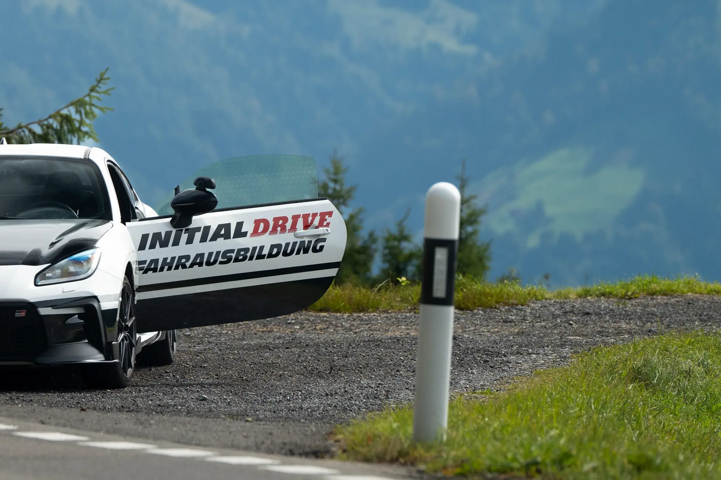 Toyota GR86 von Initial Drive mit offener Tür an einer Bergstrasse, daneben ein Leitpfosten