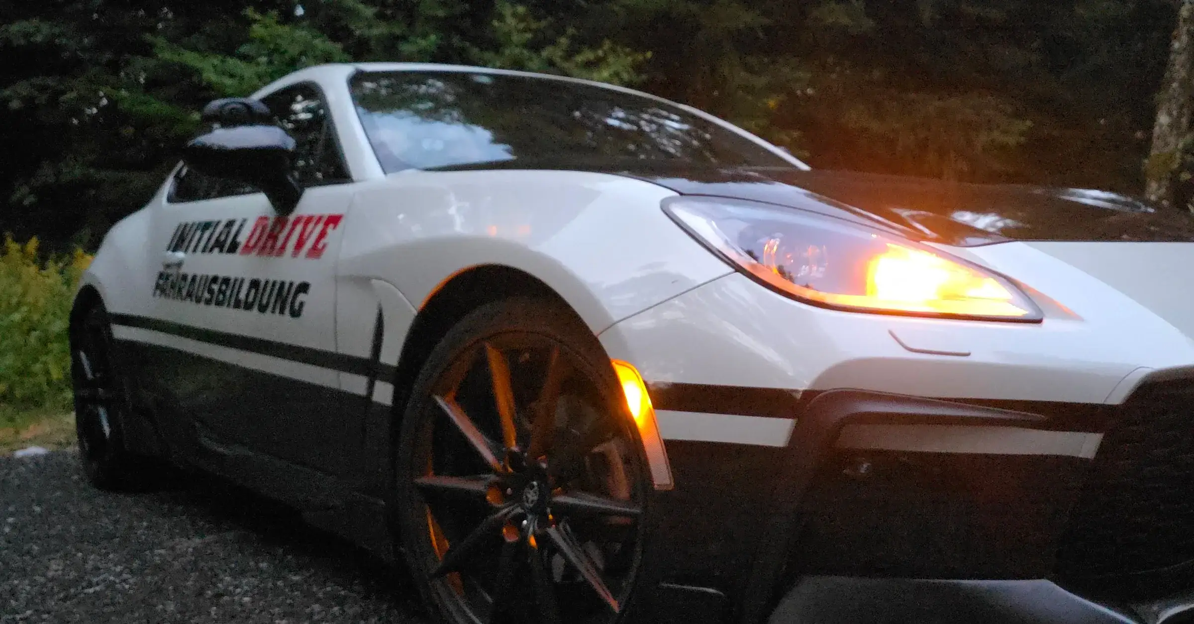 The Initial Drive Toyota GR86 at dusk with its daytime running lights on, in front of a forest