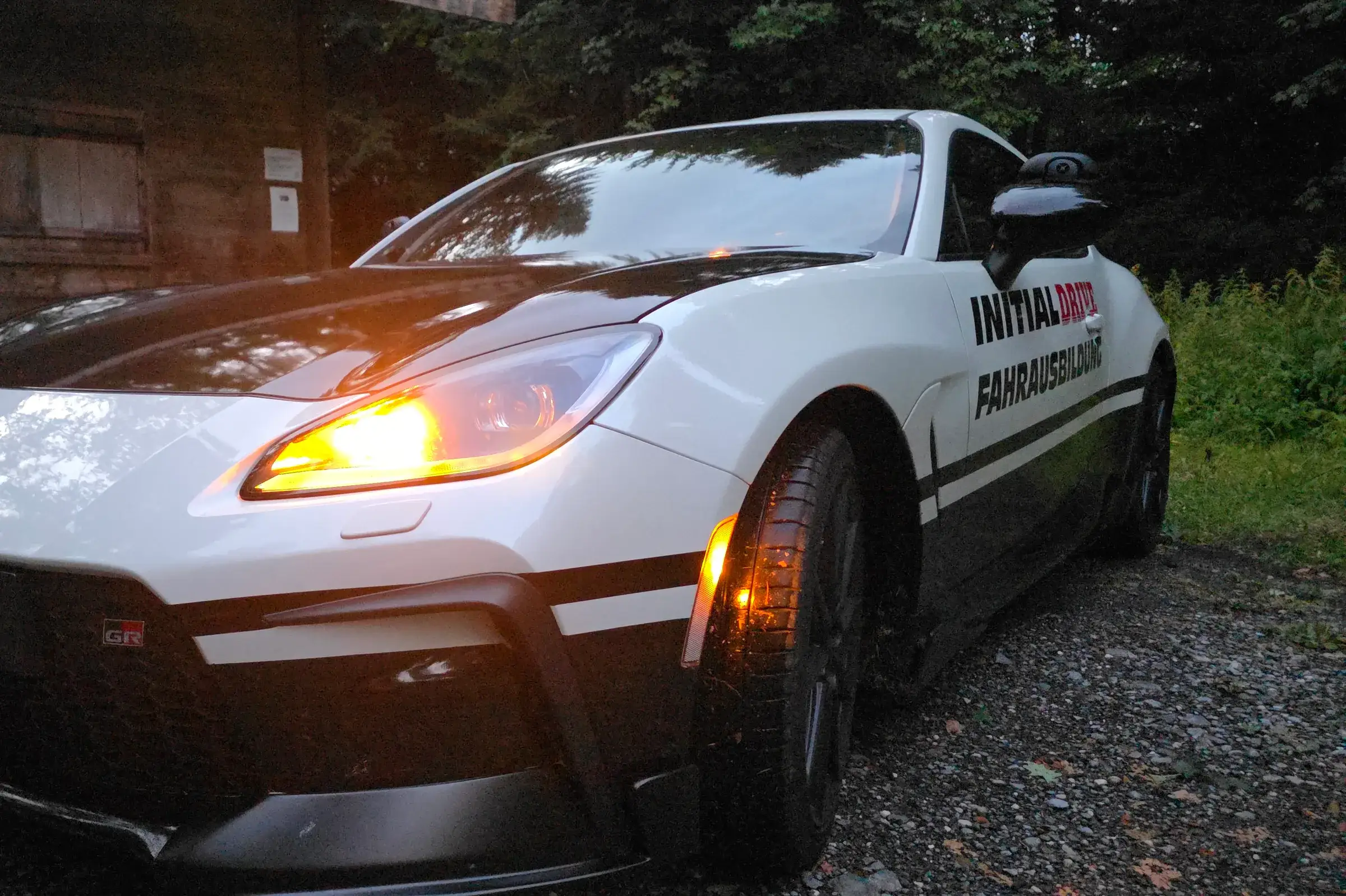 Toyota GR86 von Initial Drive mit eingeschaltetem Licht in der Dämmerung vor einer Holzhütte